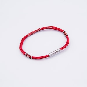 Puede incluir: Un brazalete rojo hecho de múltiples hebras de cordón con detalles en metal plateado. El brazalete tiene un cierre magnético.