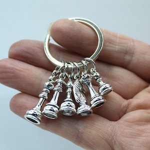 May include: A silver-coloured keyring with miniature chess pieces, including a rook, knight, bishop, queen, and pawn. The chess pieces are attached to a silver ring. The keyring is held in a hand.