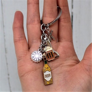 May include: Silver keychain with beer-themed charms. Charms include a bottle cap, a beer bottle with the word "BEER", a beer mug, a bottle opener, and the letter "K". The keychain is held in a hand.