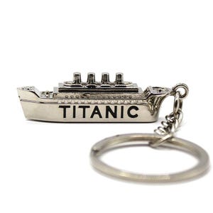 May include: A silver-coloured keychain featuring a miniature replica of the Titanic ship with the word "TITANIC" engraved on the side. The keychain has a silver-coloured metal ring.
