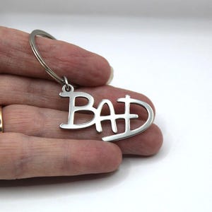 May include: A silver-toned metal keyring featuring the word BAD in a stylised, interconnected font. The letters are cut out from a single piece of polished metal, attached to a standard circular split ring for holding keys securely.