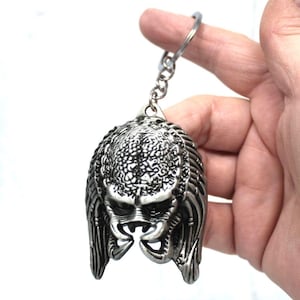 Könnte beinhalten: Ein silberner Schlüsselanhänger mit einem detaillierten Predator-Kopf mit Dreadlocks. Der Schlüsselanhänger ist an einem silbernen Metallring befestigt.