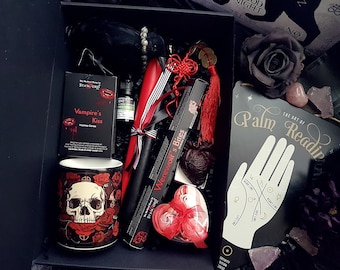 May include: A black gift box filled with various items, including a red candle, a black incense stick, a red and black skull mug, a small bottle of essential oil, a red and black incense cone, a red heart-shaped box, and a black and white palm reading guide. The box also includes a red and black tassel and a small red heart.