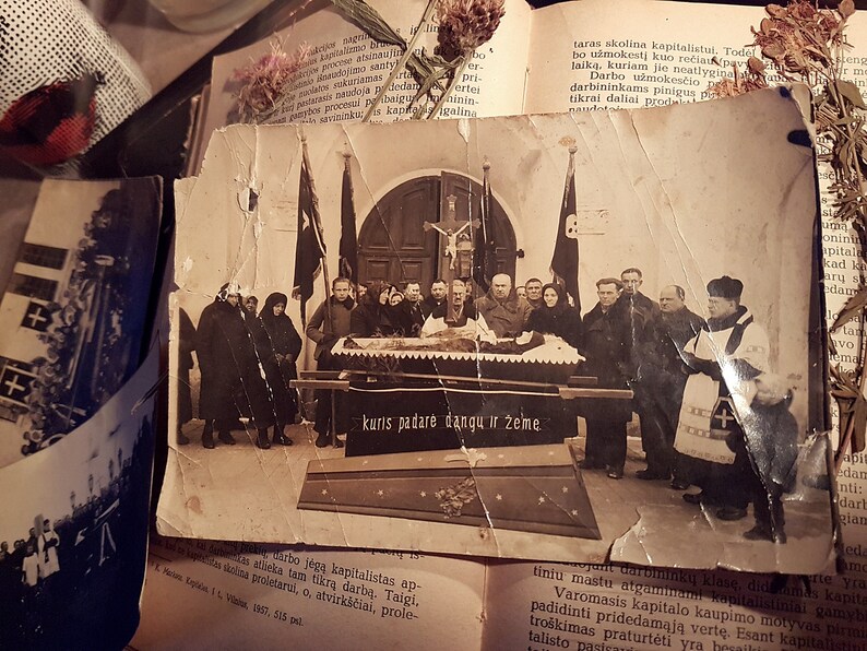 K&ouml;nnte beinhalten: Ein Schwarzwei&szlig;-Foto aus der Zeit, das eine Trauerprozession zeigt. Eine Gruppe von Menschen in dunkler Kleidung steht um einen Sarg herum. Der Text "kuris padarė dangu ir žeme" ist am unteren Rand des Bildes zu sehen.