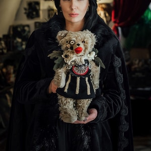 Puede incluir: Una mujer con una capa de terciopelo negro sostiene un oso de peluche vintage con una falda negra y dorada, adornada con botones, cuentas y un ojo rojo.