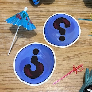 Può includere: Due adesivi circolari blu e bianchi con un punto interrogativo e una lettera j minuscola in nero su una superficie di legno.