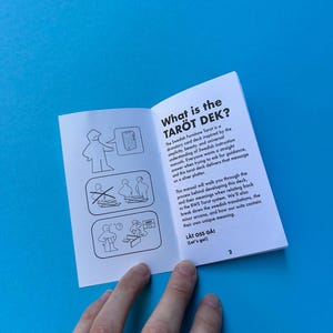 May include: An open booklet titled "What is the TAR&Ouml;T DEK?" on a blue surface. The booklet's pages feature text and illustrations. A hand is holding the booklet. The text describes a tarot card deck inspired by Swedish instruction manuals.