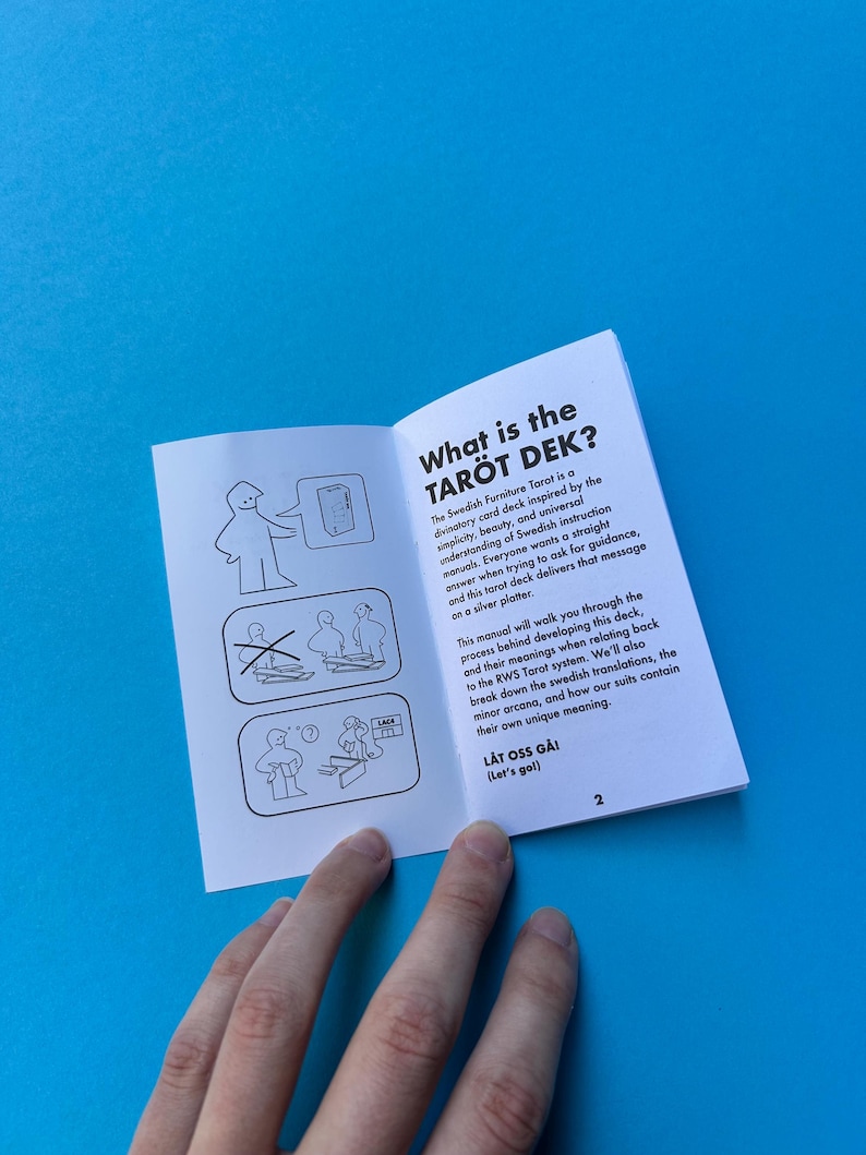 May include: An open booklet titled "What is the TAR&Ouml;T DEK?" on a blue surface. The booklet's pages feature text and illustrations. A hand is holding the booklet. The text describes a tarot card deck inspired by Swedish instruction manuals.