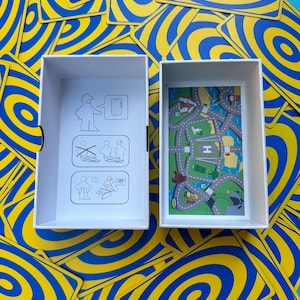 May include: A white box with a road map design and a box with instructions sits among a pile of blue and yellow swirl-patterned cards. The road map features roads, buildings, and a helipad. The cards have a concentric circle design.