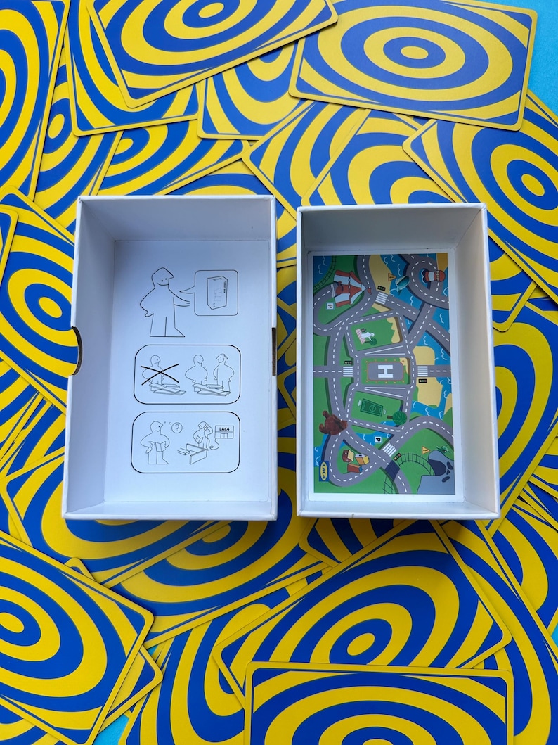 May include: A white box with a road map design and a box with instructions sits among a pile of blue and yellow swirl-patterned cards. The road map features roads, buildings, and a helipad. The cards have a concentric circle design.