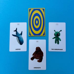 May include: A tarot card spread featuring four cards and one face-down card. The cards depict a shark, a bear, and a green alien-like figure. The face-down card has a yellow and blue concentric circle design. The cards are on a blue surface.