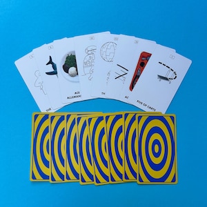 May include: A deck of playing cards with a blue and yellow concentric circle design on the back. The face-up cards feature black and white illustrations with text, including "ACE ALLEMAN!" and "FIVE OF CARTS", against a blue background.