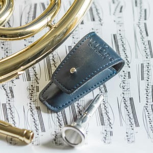 May include: A brass French horn with silver accents, a blue leather mouthpiece case, and a silver mouthpiece rest on a sheet music background. The horn has a polished finish and curved tubing.