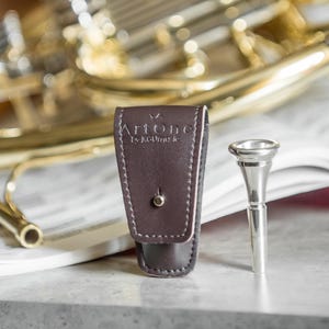 May include: A brown leather mouthpiece case with the text "ArtOne by AGUmusic" next to a silver trumpet mouthpiece. A brass instrument is in the background, resting on sheet music.