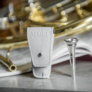 May include: A silver trumpet mouthpiece stands next to a light gray leather case with the text "ArtOne by Koumusic". The case is closed with a small silver button. The background includes a brass instrument and sheet music.