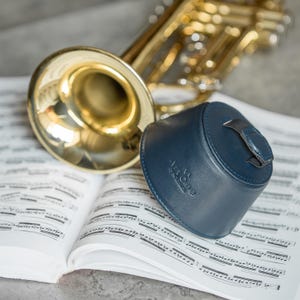 May include: A brass trumpet rests on a sheet music book next to a dark blue leather trumpet mute. The mute has a handle and the text "Art One" is visible. The background is a light gray surface.