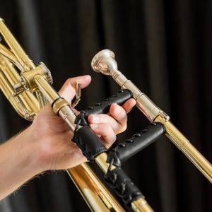 May include: A close-up of a brass trombone with a black leather grip. The trombone is held by a person's hand. The instrument has a shiny, golden finish and a silver mouthpiece. The black leather grip is stitched and provides a comfortable hold.