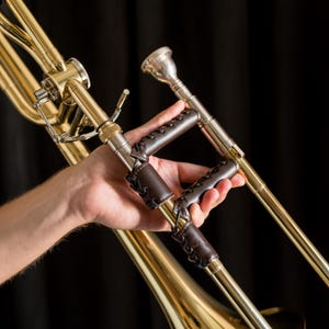 Puede incluir: Primer plano de un trombón de latón con un agarre de cuero marrón. El instrumento, parcialmente visible, muestra su acabado metálico brillante y detalles intrincados. El agarre de cuero cosido proporciona un agarre cómodo.