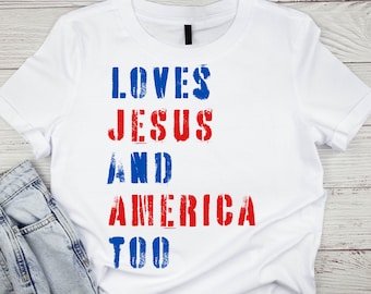 Op de afbeelding: Wit T-shirt met de tekst "LOVES JESUS AND AMERICA TOO" in rode en blauwe letters.