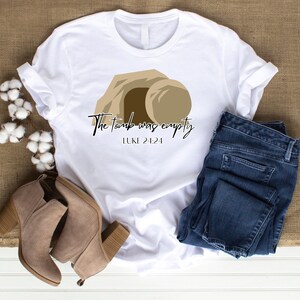 May include: White t-shirt with a brown graphic of a tomb with a stone rolled away. The text "The tomb was empty" and "LUKE 24:24" is printed on the shirt.