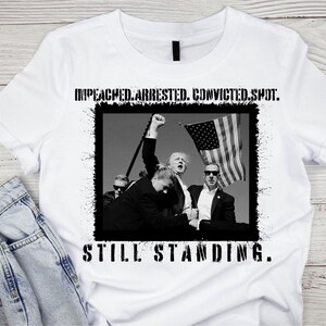Könnte beinhalten: Ein weißes T-Shirt mit einem Schwarzweißbild einer Gruppe von Menschen, die vor einer amerikanischen Flagge stehen. Der Text "IMPEACHED. ARRESTED. CONVICTED. SPOT. STILL STANDING." ist auf das Shirt gedruckt.
