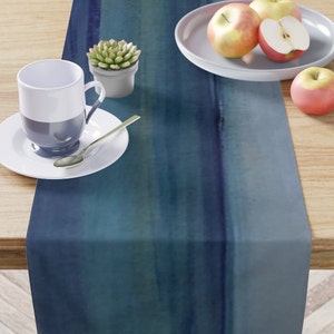 May include: A blue watercolor-style table runner with a gradient effect, transitioning from deep navy to lighter shades of teal and blue. The runner is displayed on a wooden table, with a white teacup, saucer, and spoon, along with a small succulent and apples.