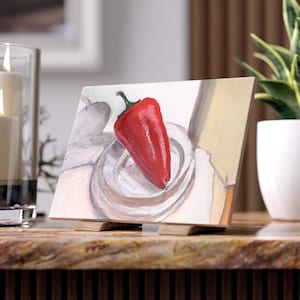 May include: A vibrant painting of a red bell pepper in a glass bowl, displayed on a wooden easel. The artwork features a realistic style with a focus on the pepper's texture and color. The painting is set against a neutral background.