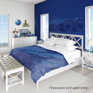 May include: A bedding set featuring a deep blue galaxy print with white star patterns on the duvet cover. The set includes white pillows and a white bed frame with a cross-patterned headboard. Text on the image reads: Pillowcases sold separately.
