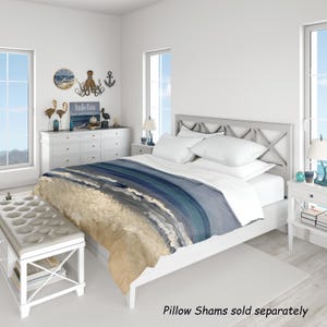 May include: A duvet cover featuring a printed design of a sandy beach meeting the ocean with shades of beige, light blue, and deep navy. The bedding is displayed on a white bed frame with white pillows, positioned in a room with white furniture.