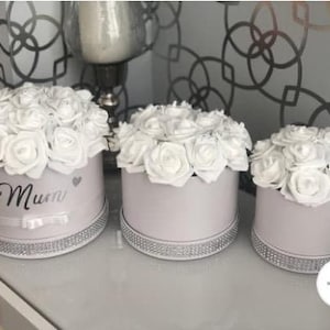 May include: Three white gift boxes with white roses and a silver trim. The box in the centre has the word "Mummy" written on it.