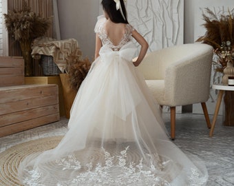 May include: A white tulle dress with a lace bodice and a large bow at the back. The dress has a long train with lace trim.