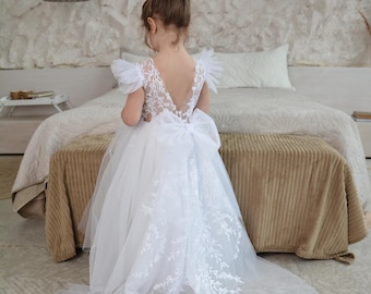 May include: A white floor-length dress featuring a V-neckline, sheer lace bodice with floral embroidery, ruffled cap sleeves, and a large bow at the waist. The skirt is made of layered tulle with an extended train adorned with white floral lace.
