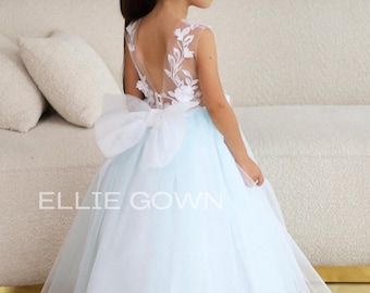 May include: A floor-length gown featuring a light blue tulle skirt and a sheer bodice with white floral embroidery. The back includes a large white bow at the waist and a deep V-neckline. The text ELLIE GOWN is printed on the front of the skirt.
