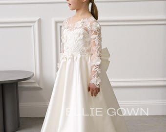 May include: A white satin flower girl dress with long sleeves and a sheer neckline. The sleeves and bodice are adorned with floral appliques. The dress has a full skirt and a large bow in the back. The text "ELLIE GOWN" is visible on the dress.