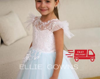 May include: A young girl wearing a light blue tulle dress with a white lace bodice and a white lace overlay. The dress has a round neckline and short sleeves. The girl is smiling and looking at the camera. The dress is for a special occasion, such as a wedding or a party.
