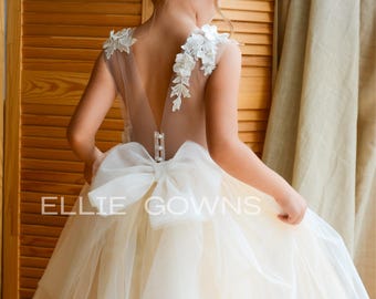 Op de afbeelding: Een vloerlange jurk met een transparante rug, witte kanten bloemapplicaties op de schouders en een grote witte tule strik in de taille. De rok is gemaakt van gelaagde gebroken witte tule. De tekst ELLIE GOWNS staat op de afbeelding gedrukt.