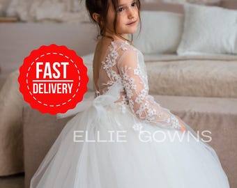 May include: A young girl wearing a white tulle dress with lace detailing and long sleeves. The dress has a bow at the back and the words "Ellie Gowns" are printed on the skirt. The girl is sitting on a bed with a white blanket. A red circle with the words "FAST DELIVERY" is in the top left corner of the image.