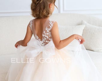 May include: A young girl wearing a white tulle dress with a lace bodice and a large white bow in the back. The dress has a sheer back and is perfect for a special occasion.