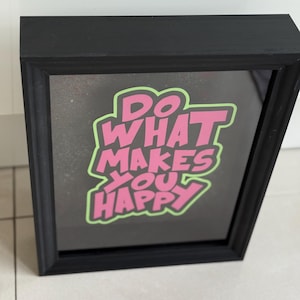 Könnte beinhalten: Schwarz gerahmter Kunstdruck mit dem Text "DO WHAT MAKES YOU HAPPY" in rosa Buchstaben mit grünem Umriss. Das Kunstwerk wird auf einem dunklen Hintergrund angezeigt.
