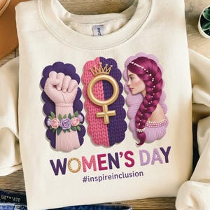 Puede incluir: Sudadera color crema con un dise&ntilde;o gr&aacute;fico del D&iacute;a de la Mujer. El dise&ntilde;o incluye un pu&ntilde;o en alto, un s&iacute;mbolo femenino con una corona y un perfil de mujer, en tonos morados, rosas y dorados. Tambi&eacute;n aparece el texto "WOMEN'S DAY" y "#inspireinclusion".
