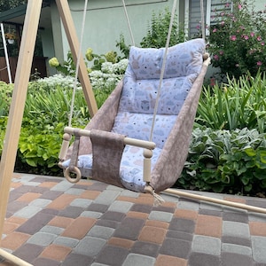 Puede incluir: Un columpio de madera con asiento y respaldo de tela beige. El asiento tiene un cojín estampado con nubes, búhos y conejos. El columpio está suspendido por cuerdas y tiene una barra de madera con un anillo de madera. El columpio está al aire libre.