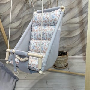 May include: A gray and white baby swing with a padded seat and a bunny pattern. The swing is made of wood and has a rope suspension system.