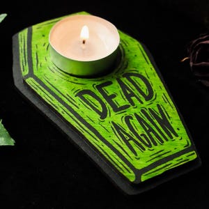 May include: A green coffin-shaped candle holder with the words "DEAD AGAIN" in black. A lit tealight candle sits in the center. The holder has a black border and is on a black surface. The artist's logo is in the bottom right corner.