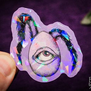 May include: A holographic sticker featuring an egg with an eye and spider legs. The egg is light purple with a detailed eye. The spider legs are black with iridescent accents. The sticker has a purple background and the artist's signature.