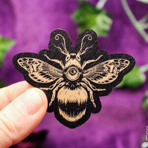 May include: A black wooden sticker with a detailed illustration of a bee with a single large eye. The bee has wings and a hairy body. The bee is in a gold color.