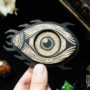 May include: A black and tan wooden eye-shaped art piece with a detailed iris and surrounding design. The art piece has a layered construction and is held by a hand. The artist's name, Fryda Gorgon Art, is visible in the lower right corner.