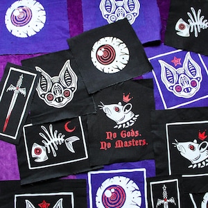 Könnte beinhalten: Eine Ansammlung schwarzer und violetter Stoffflicken mit weißen und roten grafischen Designs. Die Flicken zeigen Fledermäuse, Augen, Schwerter, Fischskelette und Ratten mit Kronen. Ein Flicken trägt die Aufschrift "No Gods. No Masters."