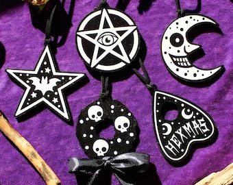 May include: A collection of five black and white hanging ornaments featuring a star with a bat, a pentagram with an eye, a crescent moon with a face, a wreath with skulls and a black ribbon bow, and a planchette shape with the text HEXMAS.