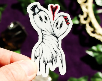 May include: A die-cut sticker featuring two white ghosts with black ink outlines, joined at the torso. One ghost wears a black top hat, the other a red bow. A small red heart is positioned between their heads, all on a white background.