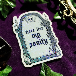 May include: A holographic sticker shaped like a tombstone featuring a skull and crossbones at the top, vines on the side, and melting candles at the base. The text "Here lies my sanity" is printed in a black gothic font on the centre surface.